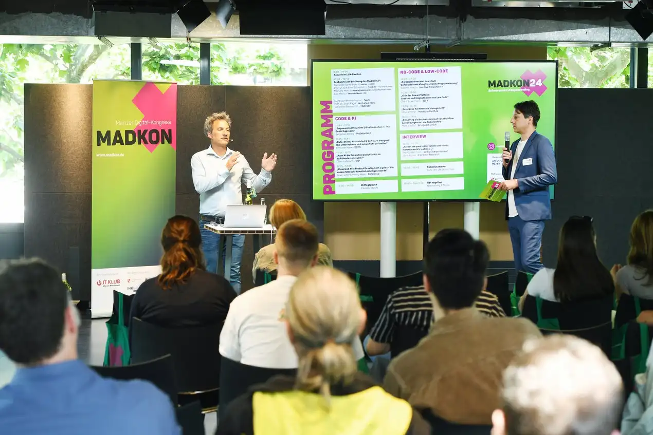 2 Personen, die vor eiem Publikum sprechen, im Hintergrund ein MADKON Banner und ein Monitor mit dem Programm der MADKON